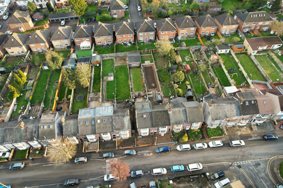 An aerial view of a residential neighbourhood showing a row of terraced houses with individual back gardens. The gardens vary in size and layout, with some featuring lush green lawns, flower beds, and