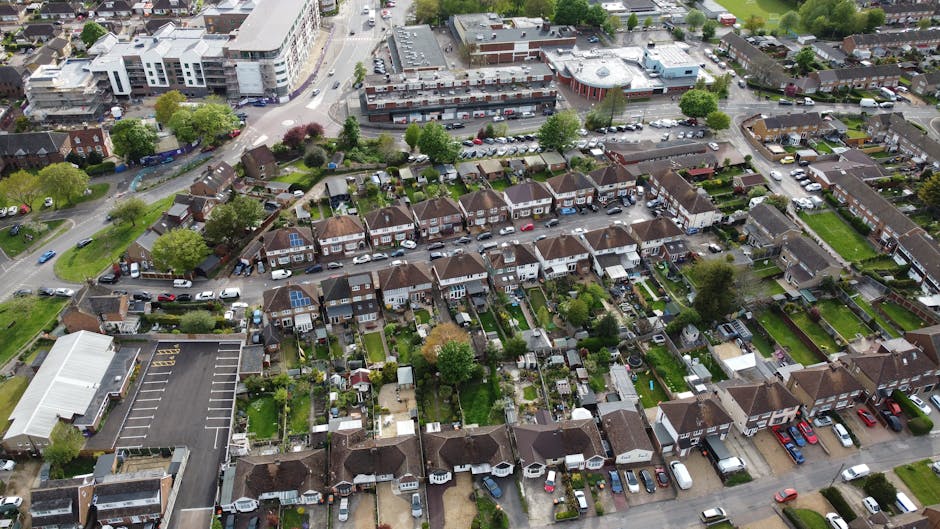 An aerial view of a residential neighborhood showing a row of small, detached houses with well-maintained gardens and lawns, surrounded by trees and shrubbery. The gardens feature green grass, flower