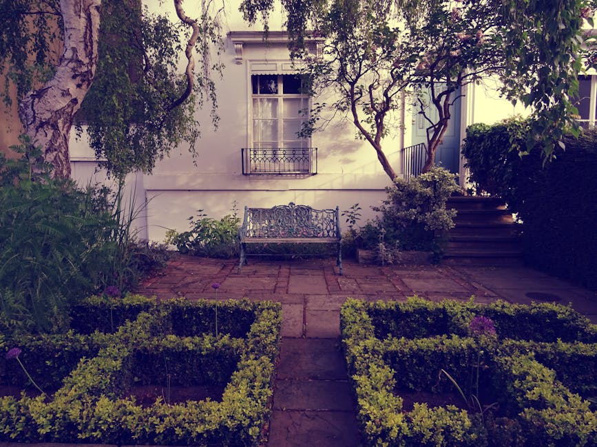 A small front garden featuring a paved patio area with a decorative metal bench positioned against a white exterior wall of a house, with a window that has a white frame and a small balcony railing. T